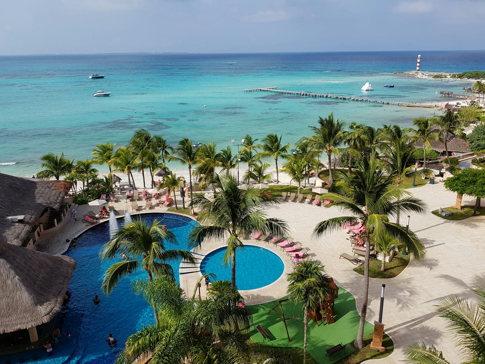 A Cancún resort seen from a balcony: blue pools and palapa bar, coconut palms, a palm-lined beach and the red-and-white Punta Cancún lighthouse on the point across turquoise water.