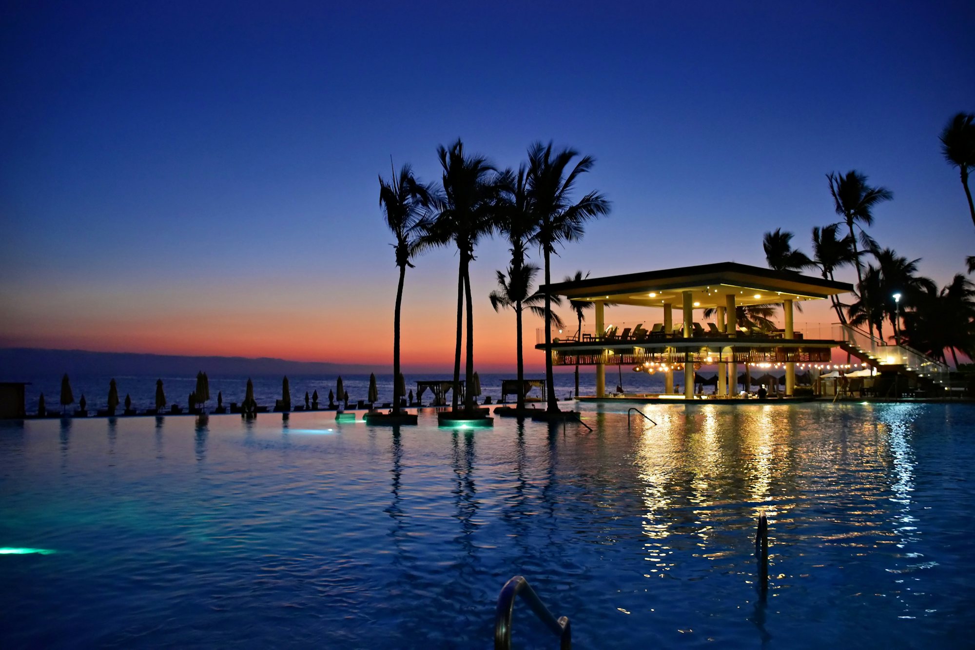 A resort infinity pool at dusk reflecting the lit pavilion and palm silhouettes, Puerto Vallarta, Mexico.