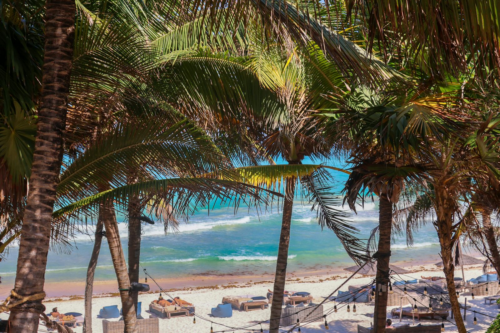 Looking through a canopy of palm fronds to a Riviera Maya beach club at Tulum: loungers and daybeds on the sand, string lights, and turquoise surf rolling in.